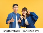 Small photo of Excited beautiful asian couple shows thumb up gesture agree to do something and collaborate against orange wall, did great job, show approval, like idea. Everything will be OK.