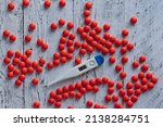 An electronic thermometer with a high temperature mark of 39.7 among many bright red pills on a wooden white table. The concept of dealing with high temperature.
