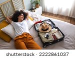 Happy woman lying down on bed next to open suitcase full of clothes, passport and hat, ready to go on vacation trip.