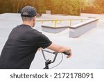 Small photo of athletic young man on bmx bike in a cap stands in a concrete bmx skate park in sunlight