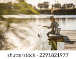 Small photo of a cute blond boy in a straw hat, jeans and rubber boots with a fishing rod on a wooden pier is fishing on a sunny, warm day