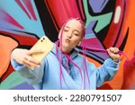 Caucasian teenage hipster girl with pink braids is using a smartphone against the background of a multicolored street wall.Summer concept.Generation Z style.Social media concept.