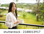 An Asia girl in white shirt is standing on balcony and holding a white cup with blur background and copy space from Thailand.