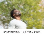 Black serious man breathing deeply fresh air in a park a sunny day with a green tree in the background