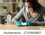 Woman with gloves and protective mask disinfecting smart phone from coronavirus sitting on a desk at night at home