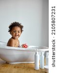 Small photo of Happy little black baby girl sitting in bath tub playing with toys in bathroom. Portrait of baby bathing in white bath full of foam, at home. everyday life. hygiene, children, lifestyle concept