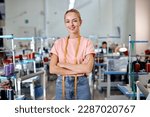 Small photo of female designer of clothes standing among sewing machines in textile factory, attractive tailor after work stand posing, looking at camera smiling with arms crossed, measuring tape for sewing on neck