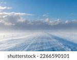 View over snow covered road in agricultural landscape in Iceland with high winds blowing snow over road creating a blur with snow clouds in distance giving way to clear blue sunny sky