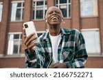 Authentic portrait of emotional  hipster guy with stylish hairstyle using mobile phone listening music, laughing outdoors. Happy African American man holding smartphone communication on the street 
