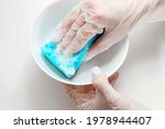 Feminine hands holding blue sponge with foam and washing white dish. Hands in transparent disposable vinyl gloves. Top view, close up, copy space 