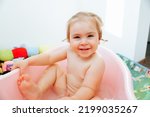 Front view looking at camera baby kid girl sits in the bath one leg raised up with her hands holding on to the bath smiling laughs. Curly hair and a happy look. Light background.