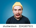 Small photo of Close up young mad angry displeased guy in yellow cap looking at camera screaming. Young Caucasian angry man yells directly into the camera. Men emotions blue background, Furious man. Stress hysteria.