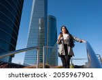 Small photo of Front view of an unsmiling adult woman standing on a step of a park staircase using phone looking away with buildings behind in a sunny day.