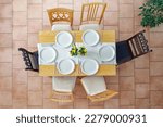 Small photo of Top view of a place setting with six white plates, cutlery and yellow daffodil flowers on a light wooden table and six different chairs on a terracotta tiled floor, selected focus