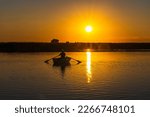 The silhouette of a 60-year-old fisherman in a wooden rowing boat with oars against the background of sunset at dusk. The yellow sun is reflected by the path on the water