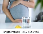 Small photo of Vitamin D capsules and glass of water and pregnant woman at home interior. Healthy fatty acids nutritional supplement for prenatal support. Omega, DHA, vitamin D, fish oil for healthy pregnancy.