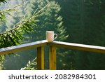 Small photo of White ceramic cup of tea, coffee beverage stands on wooden balcony railing against green coniferous forest background, tall pines, firs in woods. Breakfast in the mountains. Spruce branch in sunlight.