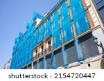 Small photo of Reconstruction of facade of a historic classical apartment building on a city street in sunny day against sky. Blue facade construction mesh covers an old red house closed for renovation wall exterior