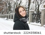 An attractive young brunette woman stands in snow-covered winter park smiles looking at camera. Portrait of pretty female girl. Path covered with white snow goes into distance. Wooden benches in a row