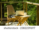 Wooden folding table, cup of a tea and laptop for remote work, chairs stand on a balcony of a country house in a green pine forest. Terrace without people. Vacations on a nature. Amazing workplace. 