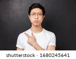 Small photo of Close-up portrait of picky, skeptical and gloomy asian guy in glasses, grimacing unsatisfied, look disappointed at camera while pointing finger left at something unpleasant and uncool