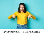 Shopping concept. Surprised cute asian girl checking out discounts, pointing fingers down and looking amazed, saying wow, standing over blue background.