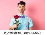 Cute and funny man waiting for kiss from lover on Valentines day, holding beautiful red rose for girlfriend, standing over pink background