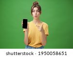 Small photo of Portrait of intrigued woman cannot wait use new smartphone holding phone and pointing at cellphone screen folding lips with excitement and interest presenting new device model in store over green wall