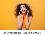 Waist-up shot of attractive silly curly-haired european female in cropped top using wireless earphones touching earbuds and gazing at upper right corner delighted and carefree over orange wall