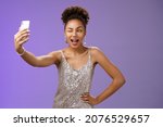 Small photo of Stylish sassy african american b-day girl taking selfie new elegant silver shiny dress extend arm holding smartphone posing winking display screen amused having fun smiling broadly, blue background