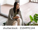 Smiling asian woman looking outside window with dreamy face, drinking morning coffee and sitting near her home patio