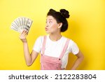 Small photo of Shopping. Silly korean girl with glam makeup, pucker lips and looking at dollar bills, wasting money, standing on yellow background