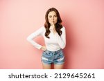 Small photo of Glamour stylish brunette girl, shush, silence you, press finger to lips and winking, sharing secret, hushing, be quiet sign, standing over pink background