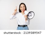 Smiling happy asian girl talking in megaphone and pointing up, announcing discount promo, showing advertisement on top, standing over white background