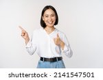 Image of young asian business woman, smiling while pointing finger left and showing thumbs up, recommending product, praise, standing over white background