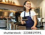 Small photo of Portrait of beautiful asian girl, student working part-time in cafe, holding cup of coffee, made order, looking for client, wearing uniform.