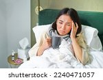 Portrait of asian woman with headache, catching col, staying on sick leave at home, lying in bed, drinking hot tea, having flu.