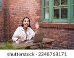 Friendly asian woman sitting with laptop, waving at you, saying hello, hi gesture, greeting you while working with computer.