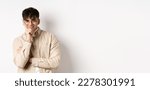 Small photo of Cunning young man watching something interesting happen, lean face on arm and smiling devious, looking at camera intrigued, standing on white background.