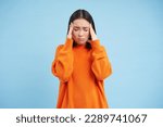 Frustrated asian woman holds hands on head, has headache, looks miserable, cant handle pressure, feels distressed, stands over blue background. Mental health concept.