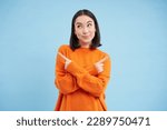 Small photo of Decisions. Young asian woman points sideways, shows left and right with indecisive smiling face, choosing between two options, stands over blue background.