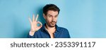 Small photo of Image of silly young man pucker lips and show okay gesture, praise good thing, compliment excellent choice, standing on blue background.