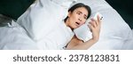 Small photo of Portrait of asian woman waking up in bed, looking shocked at mobile phone, realise she overslept.