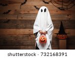 Halloween Concept - little white ghost with halloween pumpkin candy jar doing trick or treat with curved pumpkins over bats and spider web on Wooden studio background.