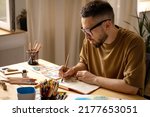 A young artist makes a brush sketch at his desk. Portrait of a creative person in his studio. Creative studio, lifestyle, the process of creating a work of art, the search for inspiration.