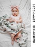 Small photo of Astonished infant in trendy clothes and bow lying on soft bed under coverlet with floral ornament. Stylish baby with muslin swaddle blanket on bed