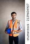 Small photo of An engineer of Middle Eastern descent smiling,handsome, holding a hard hat, wearing a reflective vest.With a background on the wall. Portraits about industry, construction, workers, readiness to work
