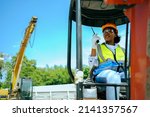 Small photo of A woman worker driving a backhoe to dig a hole in a construction site holding a walkie talkie.African American female engineer wearing a hard hat and vest. cute female with black skin gender equality.