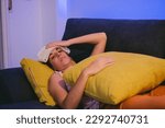 Small photo of Sick young Latin woman lying on a sofa with a towel on her head to bring down her fever while hugging a cushion.