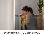 A woman covered with a blanket is freezing because of an energetic crisis at home and trying to warm sitting by the radiator.